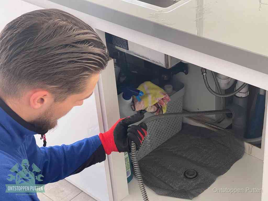 Een monteur voert een flexibele ontstoppingsveer in onder het keukenkastje, langs de sifon, om de gootsteen te ontstoppen in Putten