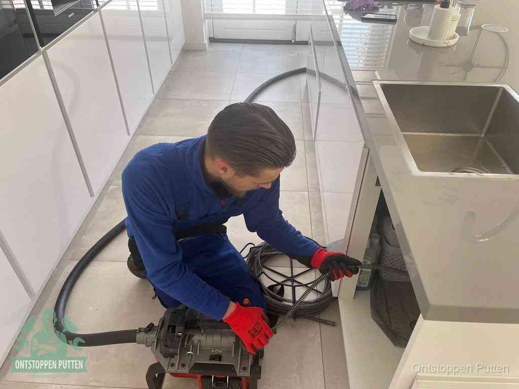 Een monteur ontstopt de keukenafvoer onder de spoelbak met een elektrische veermachine in Putten