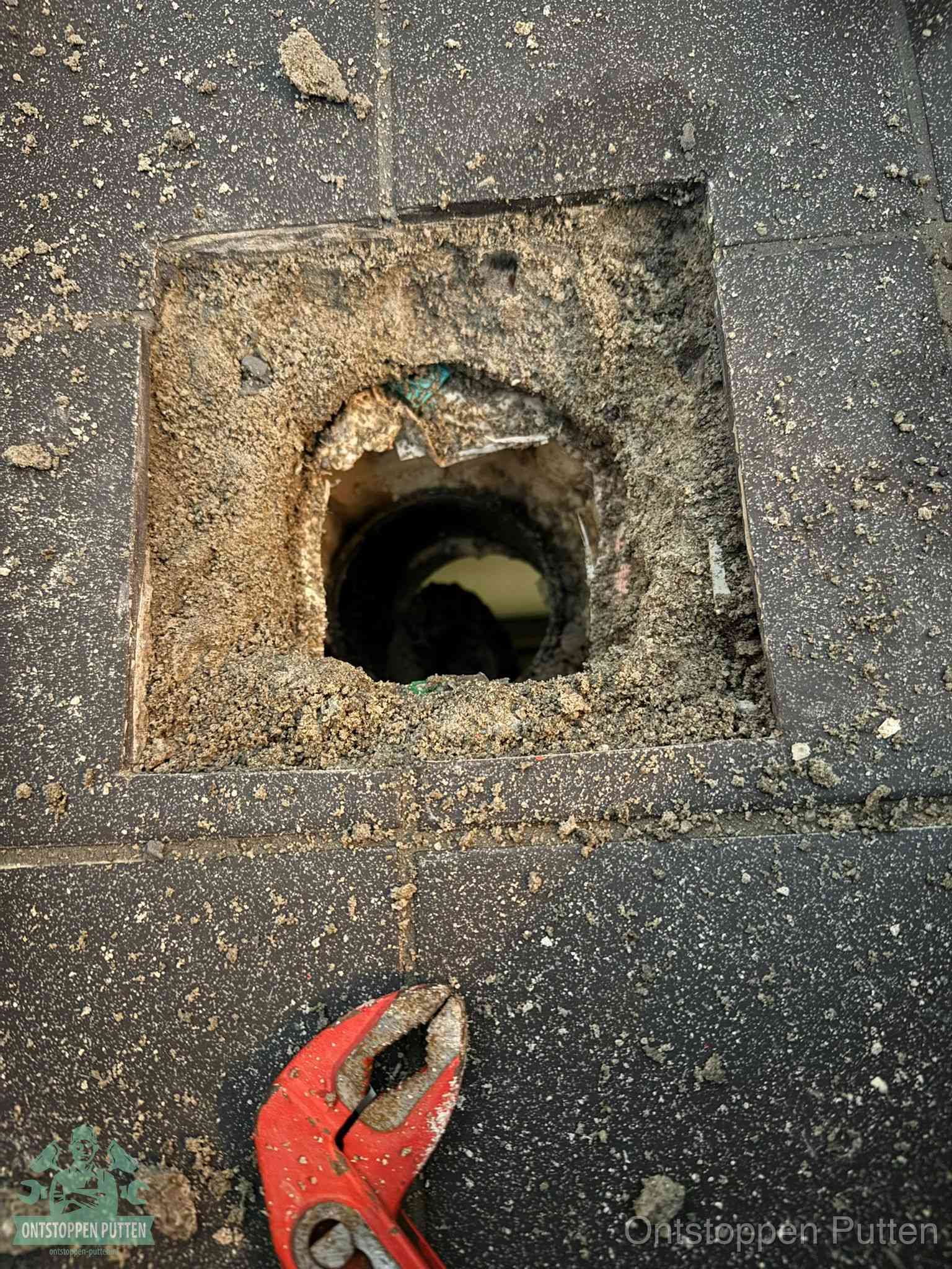 Een geopend rioolputje in een betontegelvloer met losgemaakt rooster en een zichtbare ronde afvoerbuis, het achterhalen waar de afvoerverstopping zit in Putten