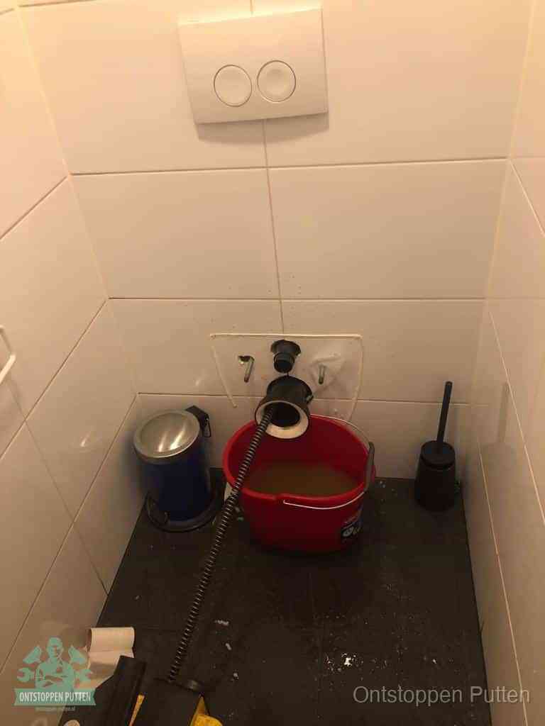Een ontstopte wand-wc met verwijderde toiletpot waar een ontstoppingsveer in de wanduitgang verdwijnt, het achterhalen waar de verstopping zit in Putten