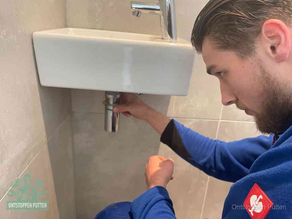 Een monteur draait met de hand de zwanenhalssifon onder een fonteinwasbak in de toiletruimte los om de bron van rioollucht in Putten te vinden