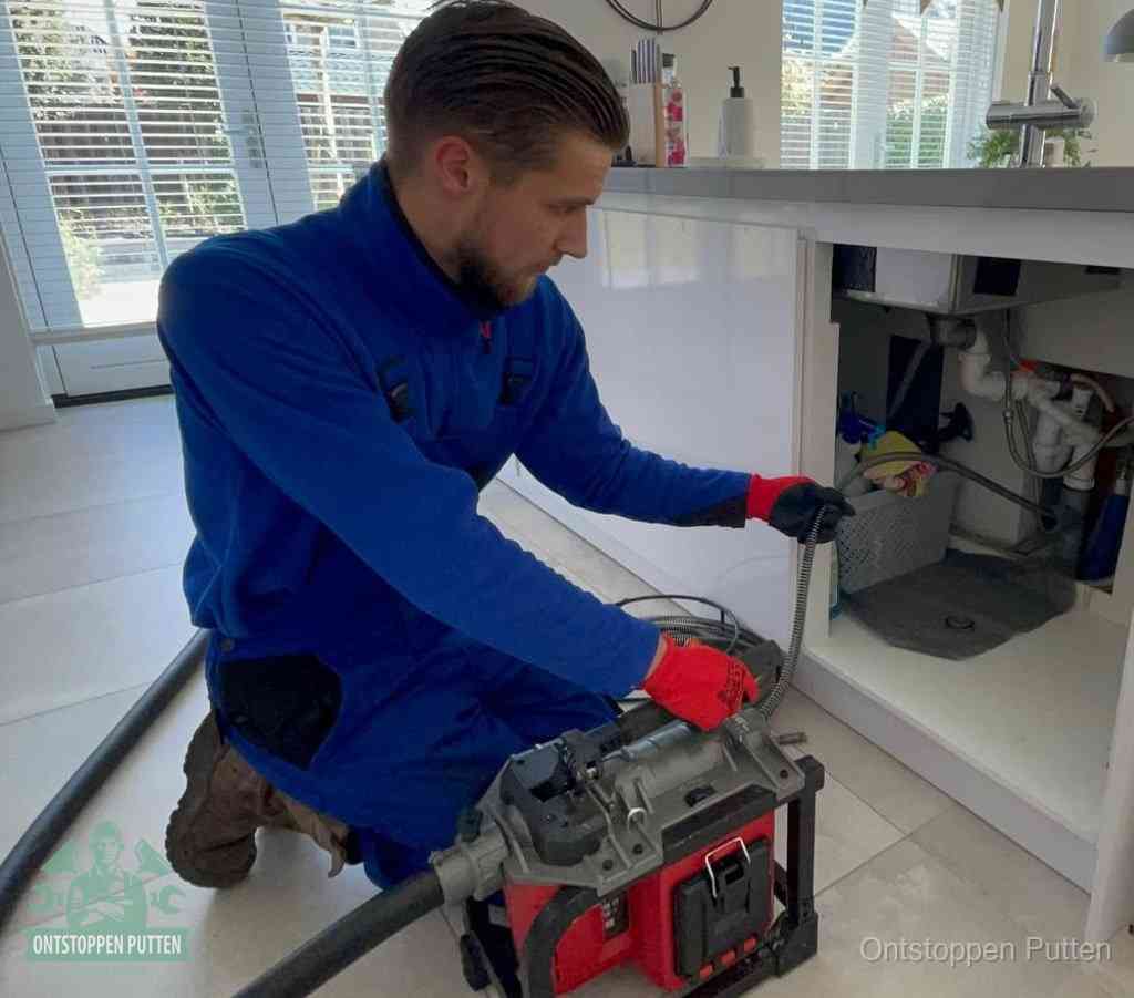 Een monteur met rode handschoenen knielt in een keuken en voert een elektrische ontstoppingsveer in tijdens het professioneel afvoer ontstoppen in Putten